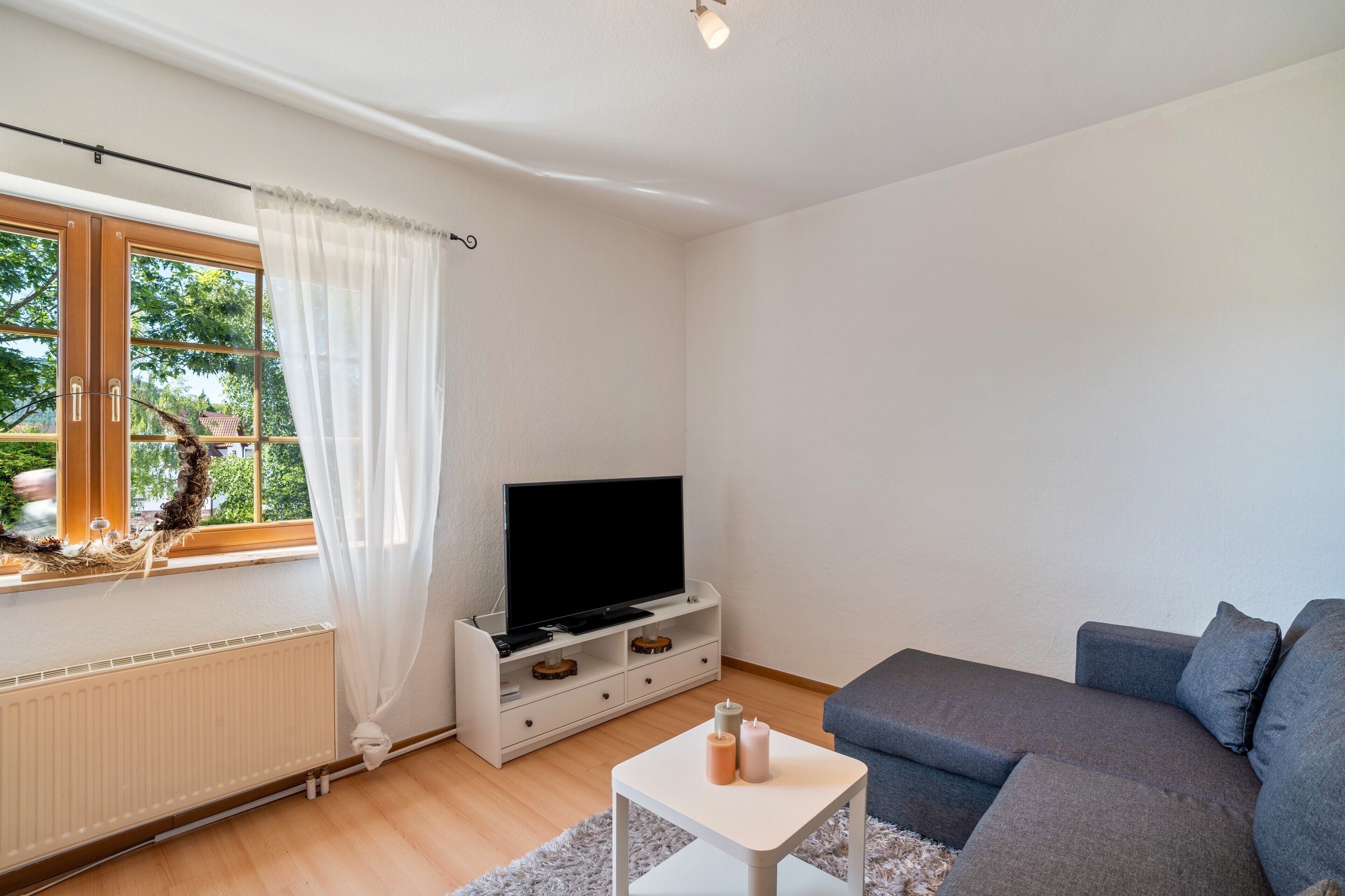 Un salon moderne avec un téléviseur à écran plat posé sur un meuble blanc, un canapé gris et une fenêtre agrémentée de rideaux blancs.