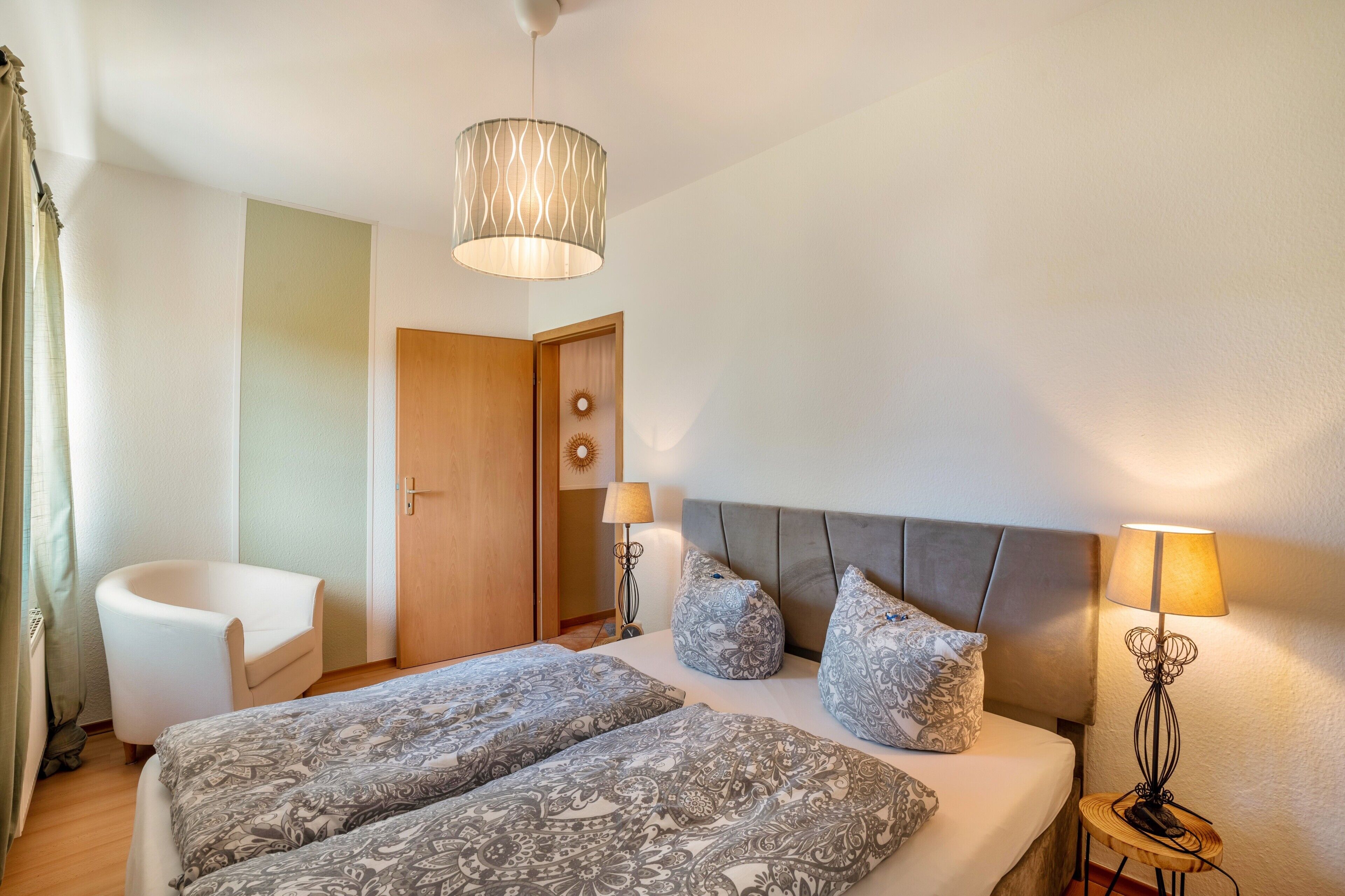 Una habitación de hotel con cama, dos lámparas de noche, una silla, una puerta y una lámpara colgante.