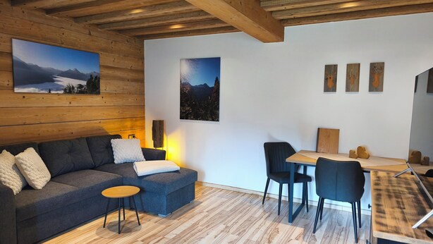 Ein modernes Interieur mit Holzbalkendecke, einer Couch, einem kleinen Tisch, einer Essecke und Wandbildern.