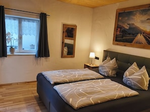 A bedroom with a bed, pillows, a nightstand with a lamp, a window with curtains, a mirror, and a painting on the wall.