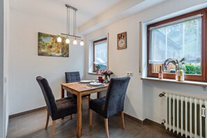 Un coin repas avec une table en bois, quatre chaises, un tableau, une horloge et une fenêtre donnant sur une maison.