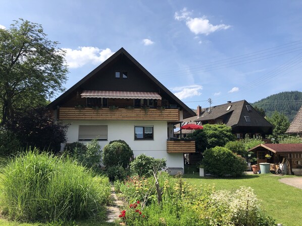 Une maison à deux étages, dotée d’un toit en pente, d’un balcon avec un parasol rouge et d’un jardin où poussent diverses plantes et fleurs.