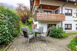 Une maison à deux étages avec un balcon, une terrasse dallée avec une table et des chaises, et un jardin avec différentes plantes.