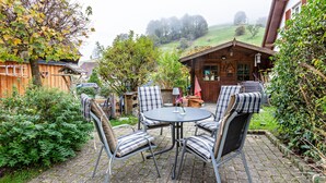 A patio with a round table and chairs, surrounded by greenery and a wooden building.