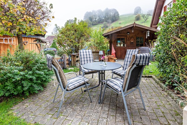 Une terrasse avec une table ronde et des chaises, entourée de verdure et bordée d’un bâtiment en bois.