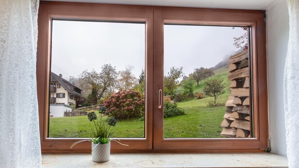 A window with a view of a green landscape, a stone wall, and a building in the background.