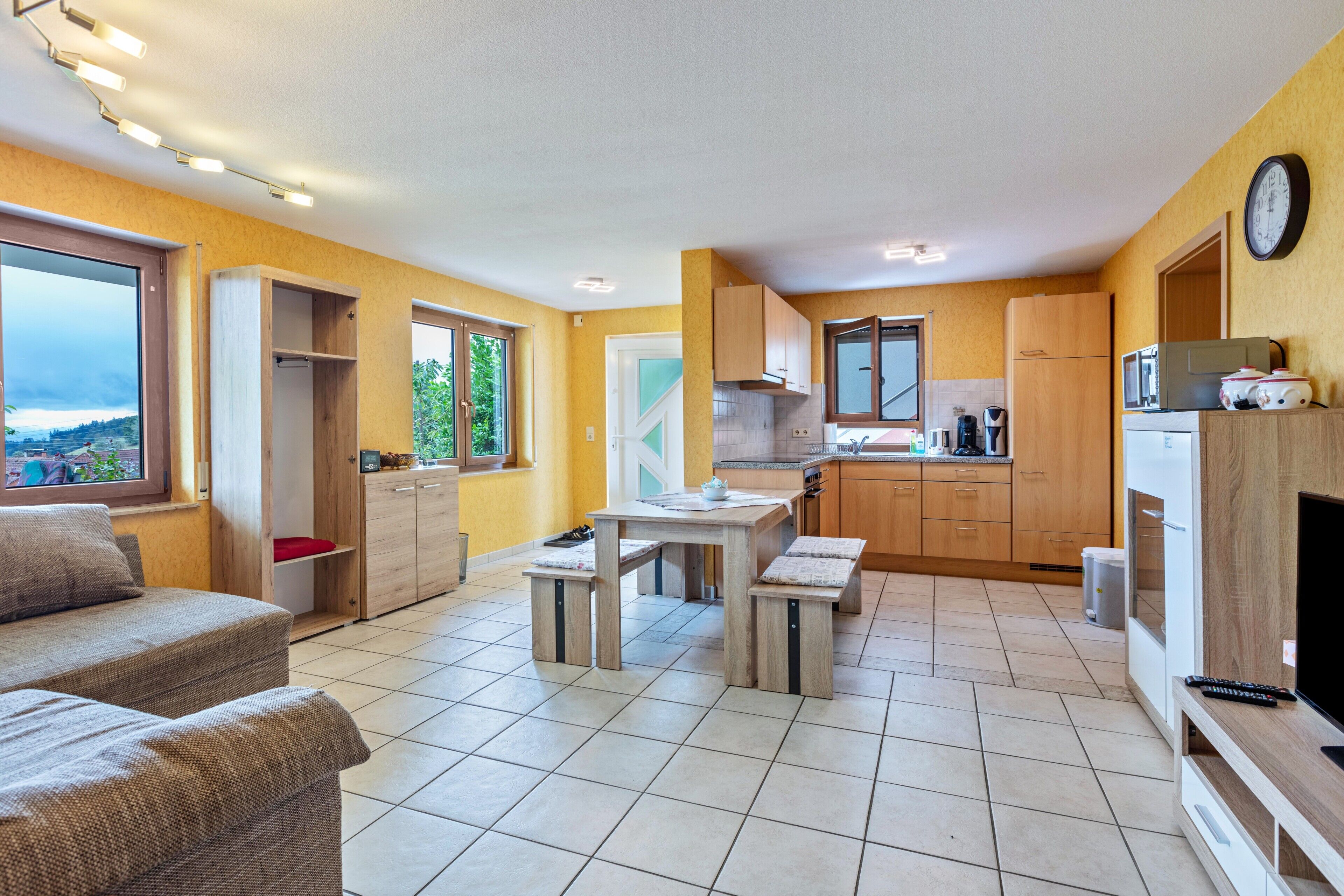 A compact living space with a sofa, a dining table, a kitchen area with wooden cabinets, and a large window offering a view.