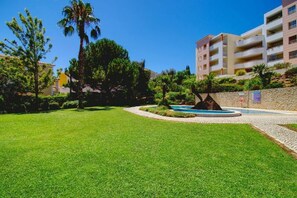 Un área verde bien cuidada con una fuente circular de agua, rodeada de árboles y un edificio con balcones.