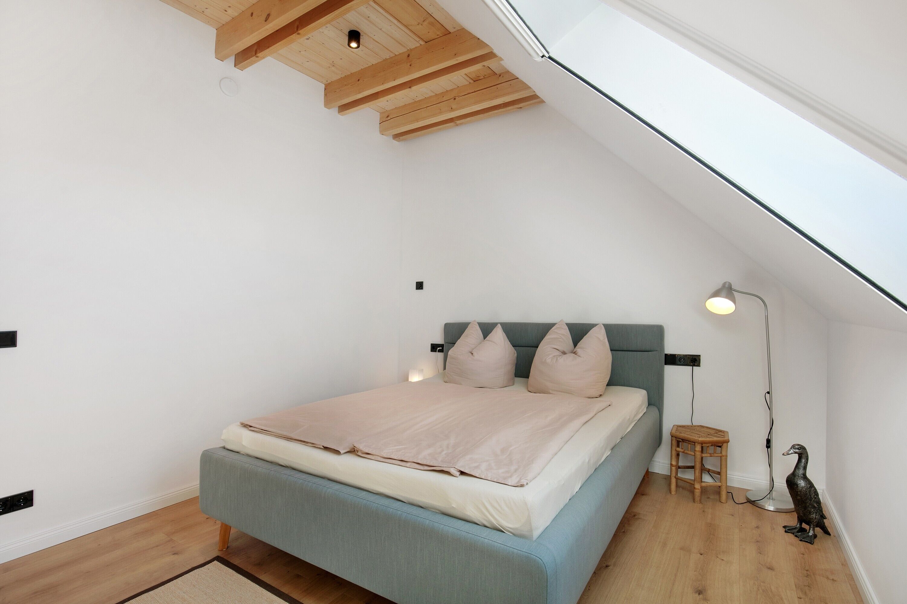 A bedroom with a sloped ceiling, a bed with a light blue headboard, a small stool, and a floor lamp.