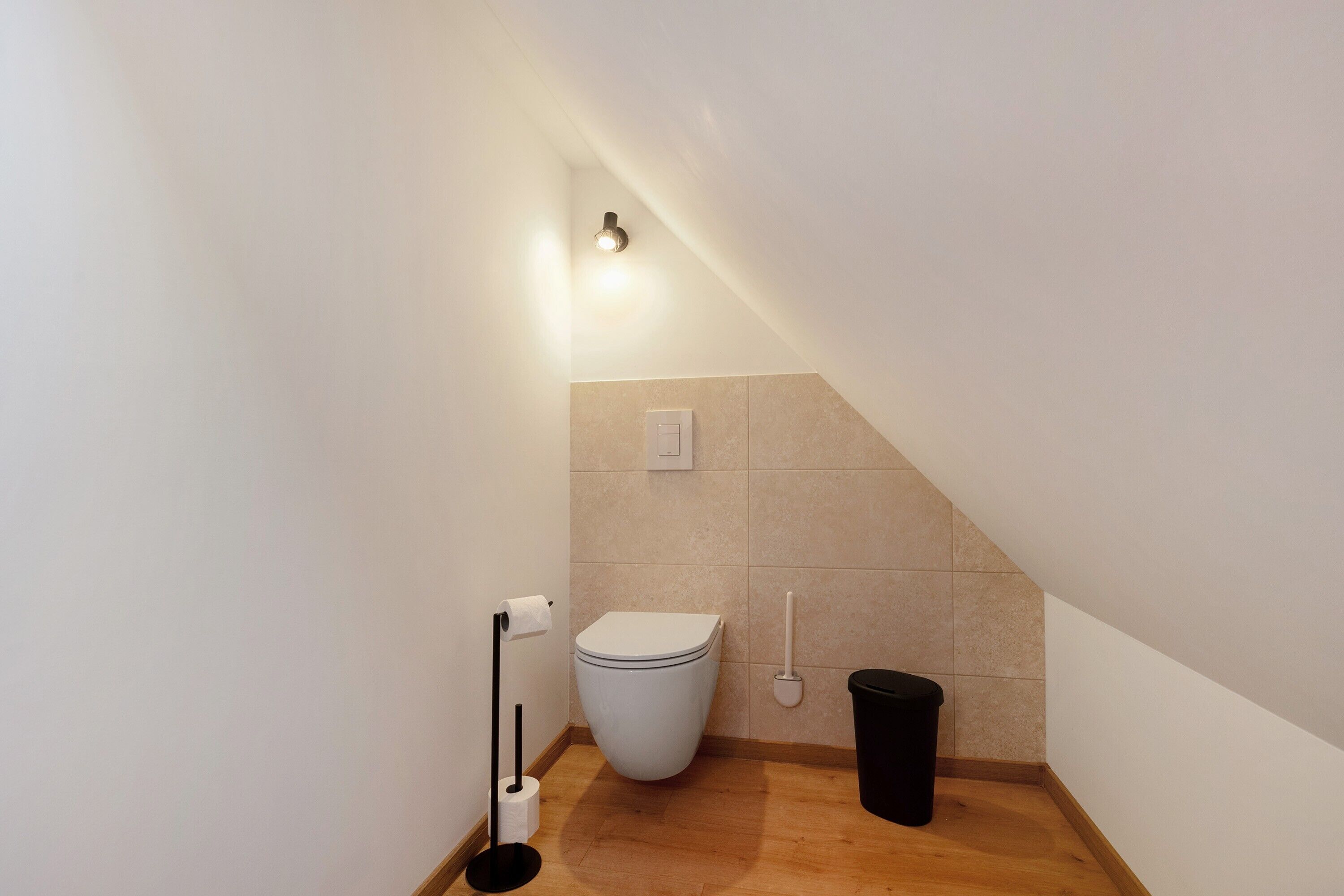 A small, modern bathroom with a toilet, a trash bin, a toilet paper holder, and a wall-mounted light fixture.