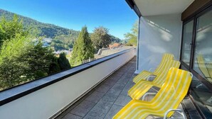 Un balcone con poltrone da relax a righe gialle, vista libera su un pendio boscoso e un edificio moderno sullo sfondo.