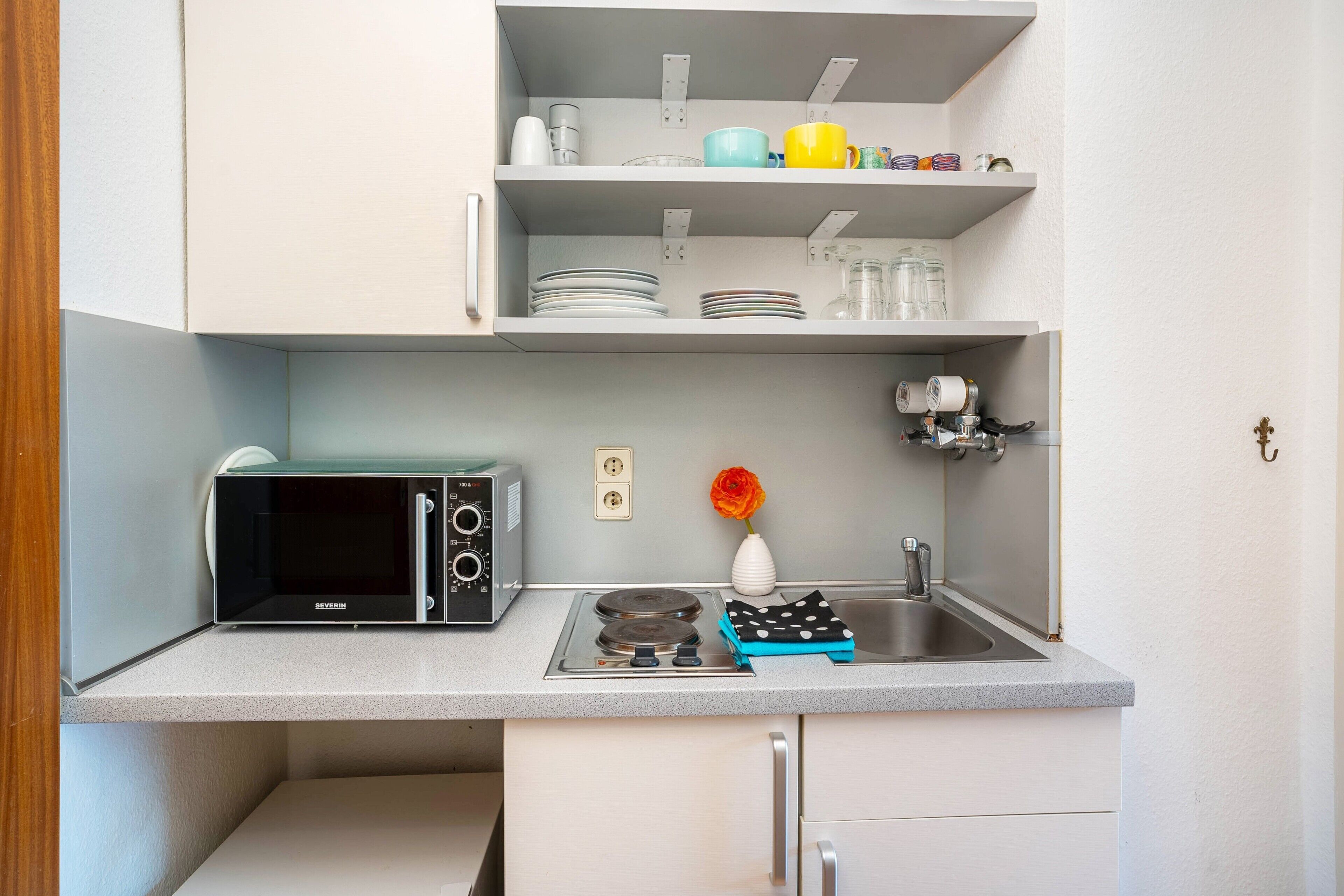 A compact kitchen with a microwave, stove, sink, and shelves with dishes and a vase.