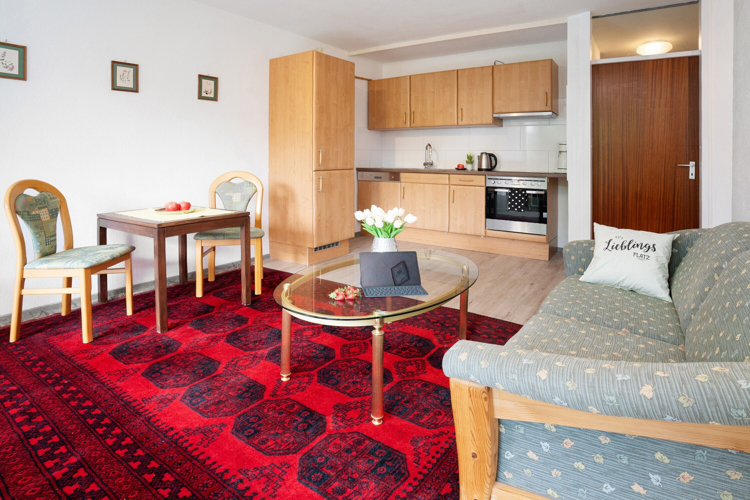 Un salon comprenant un canapé, une table basse en verre, une table à manger avec des chaises, un coin cuisine et un tapis rouge à motifs.