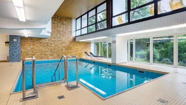 An indoor swimming pool with a tiled floor, a brick wall, and a view of trees through large windows. - Apartment No. 19 with Shared Garden, Balcony and Wi-Fi (Bad Herrenalb)