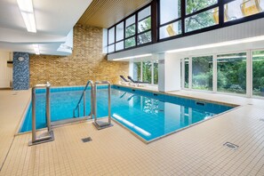En inomhuspool med ett kaklat golv, en tegelvägg och utsikt över träd genom stora fönster.