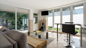 Una sala de estar moderna con una mesa de centro de vidrio, un sofá, un televisor montado en la pared y un amplio ventanal corredero que da a un espacio exterior.