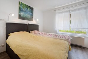 Um quarto com cama, janela com cortinas, um quadro na parede e abajur.