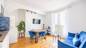 Una sala de estar moderna con piso de madera, un sofá azul, una mesa de comedor con sillas, un televisor en la pared y un ventanal con cortinas.
