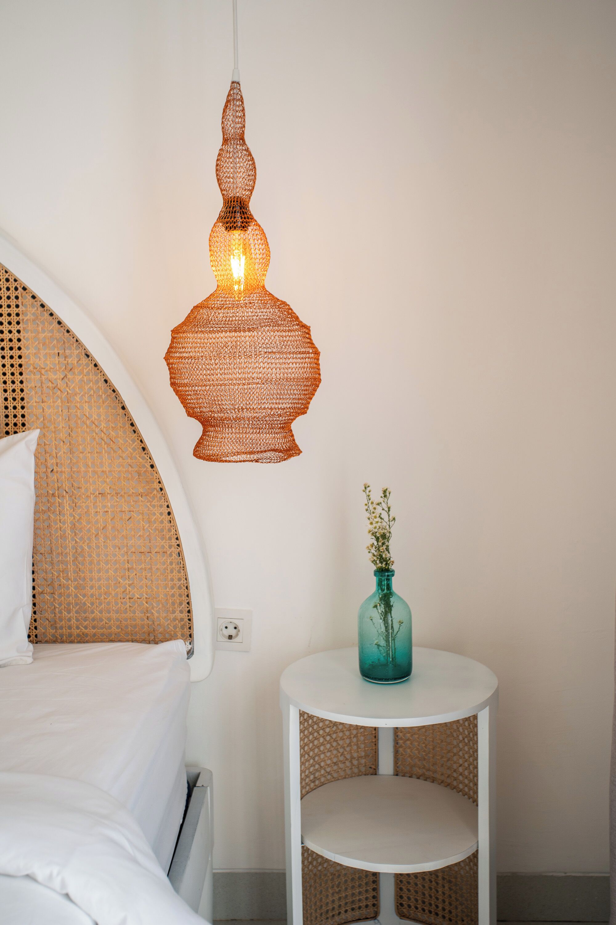 Ein Schlafzimmer mit einem gewobenen Kopfteil, einer hängenden Leuchte, einem kleinen runden Tisch mit einer Vase und einem weißen Nachttisch.