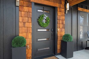 A modern entrance with a dark door, a wreath, and two potted plants.