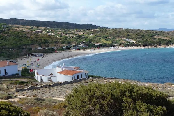 A coastal area with a beach, a white building with a red roof, and a hilly landscape in the background.