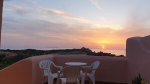 Ein Balkon mit weißen Plastikstühlen und einem Tisch, mit Blick auf einen Sonnenuntergang und eine entfernte Hügellandschaft.