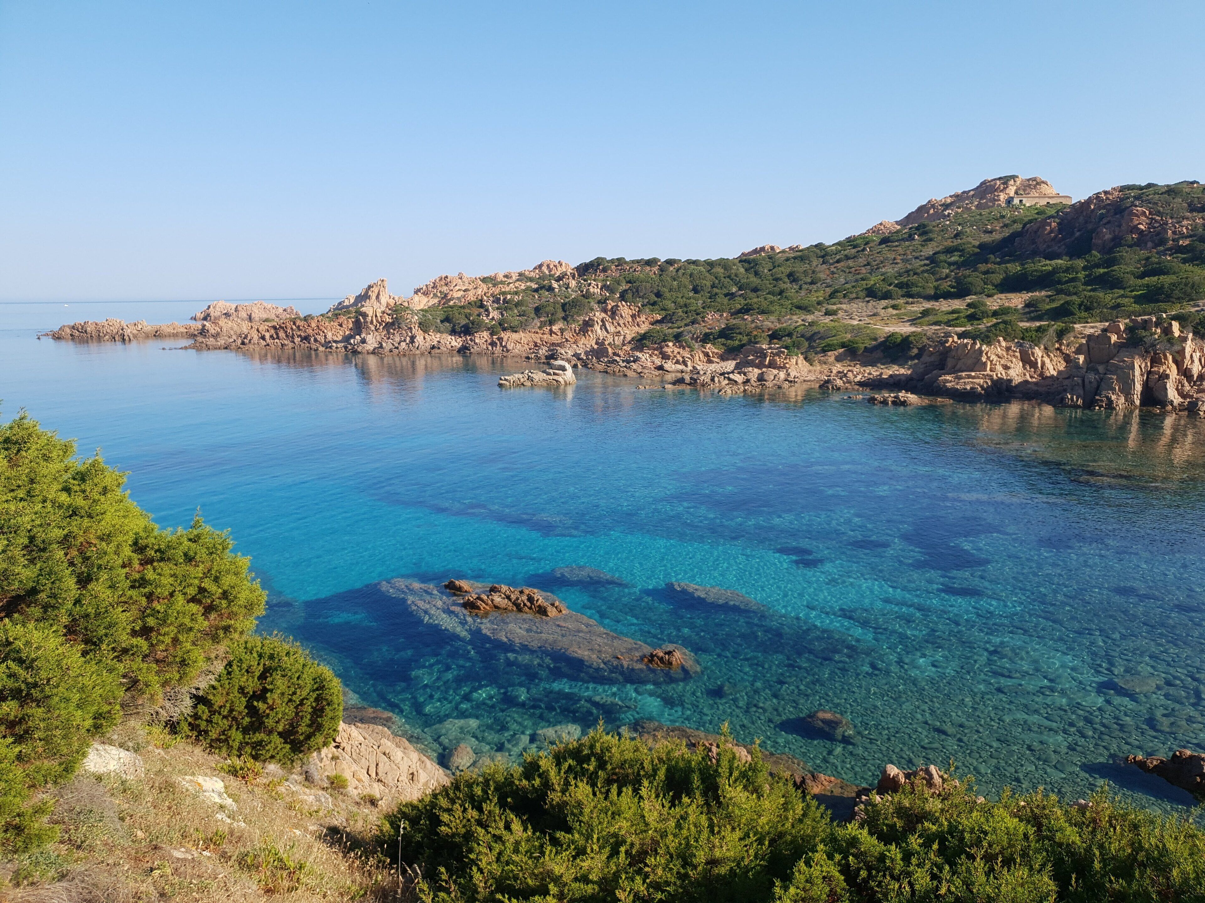 A coastal landscape with clear blue waters, rocky terrain, and greenery.