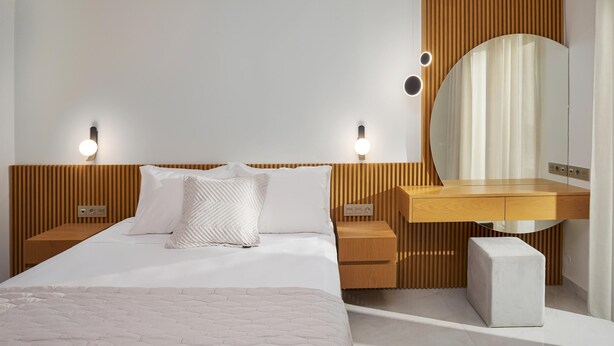 Una habitación de hotel moderna con una cama grande, cabecera de madera, mesitas de noche con lámparas, tocador con espejo y un pequeño banco blanco.