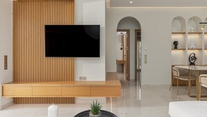A modern living room with a flat-screen TV mounted on a wooden wall, a round coffee table with candles and a plant, and a dining area with a round table and chairs. - Adore Gold Natural Retreat by Estia (Mylopotamos)