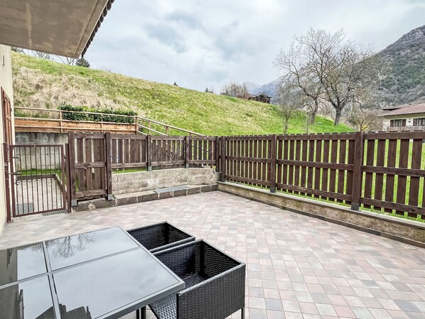 A patio with a glass table and wicker chairs, surrounded by a wooden fence and a stone wall. - Valentina by Interhome (Lago di Ledro)