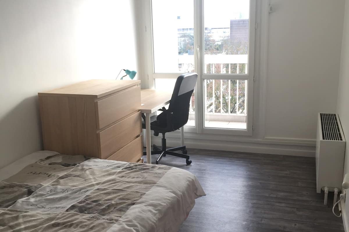 A bedroom with a bed, a wooden dresser, a desk with a chair, a radiator, and a balcony with a view of buildings.