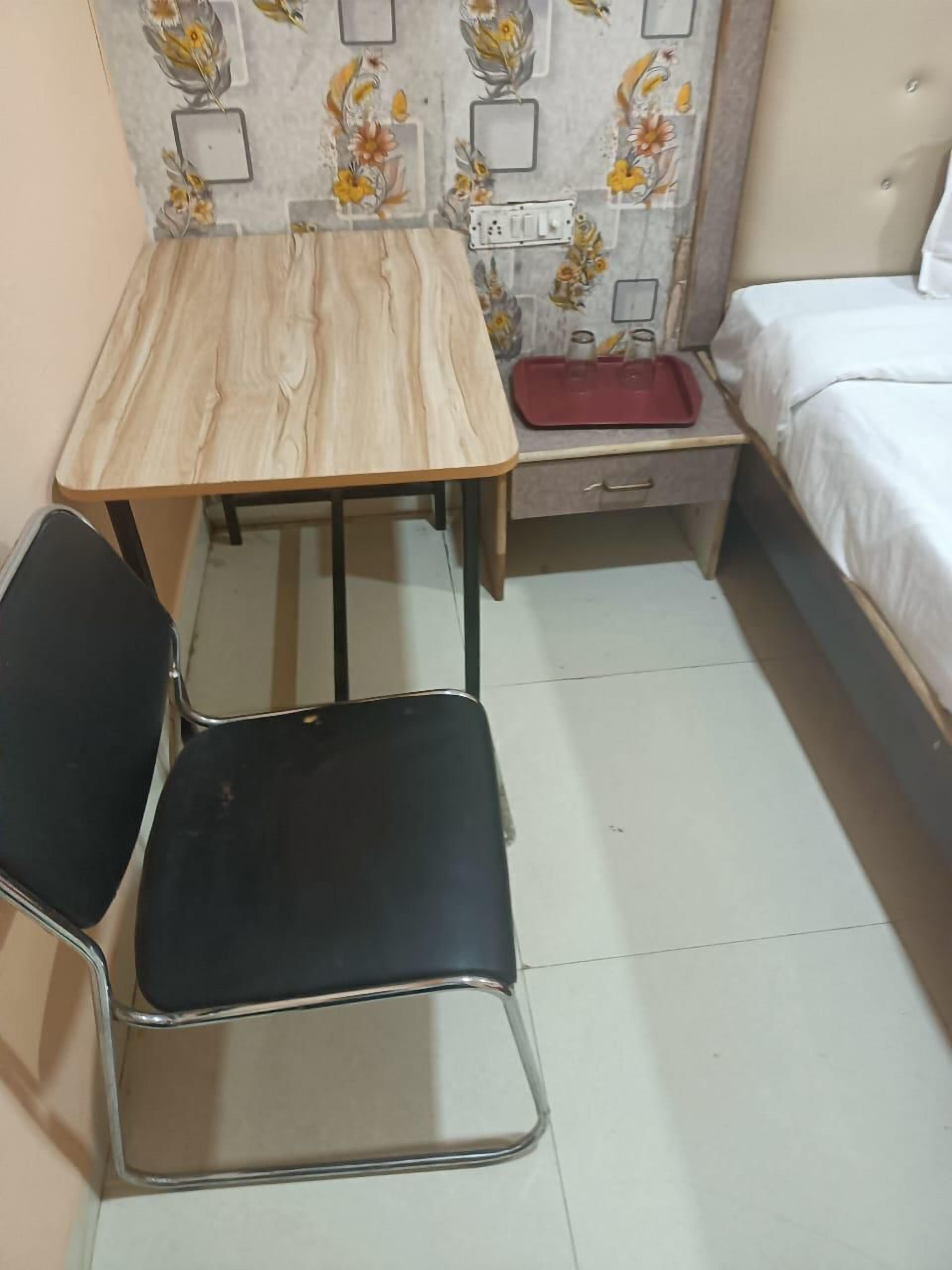 A small room with a wooden table, a black chair, a bed, and a nightstand with a red tray.