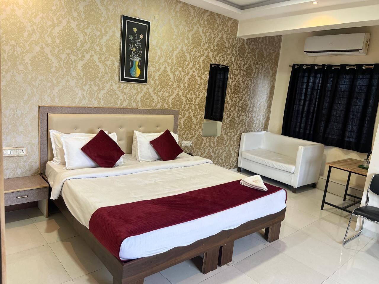 A hotel room with a large bed, a white sofa, a wooden nightstand, and an air conditioning unit.