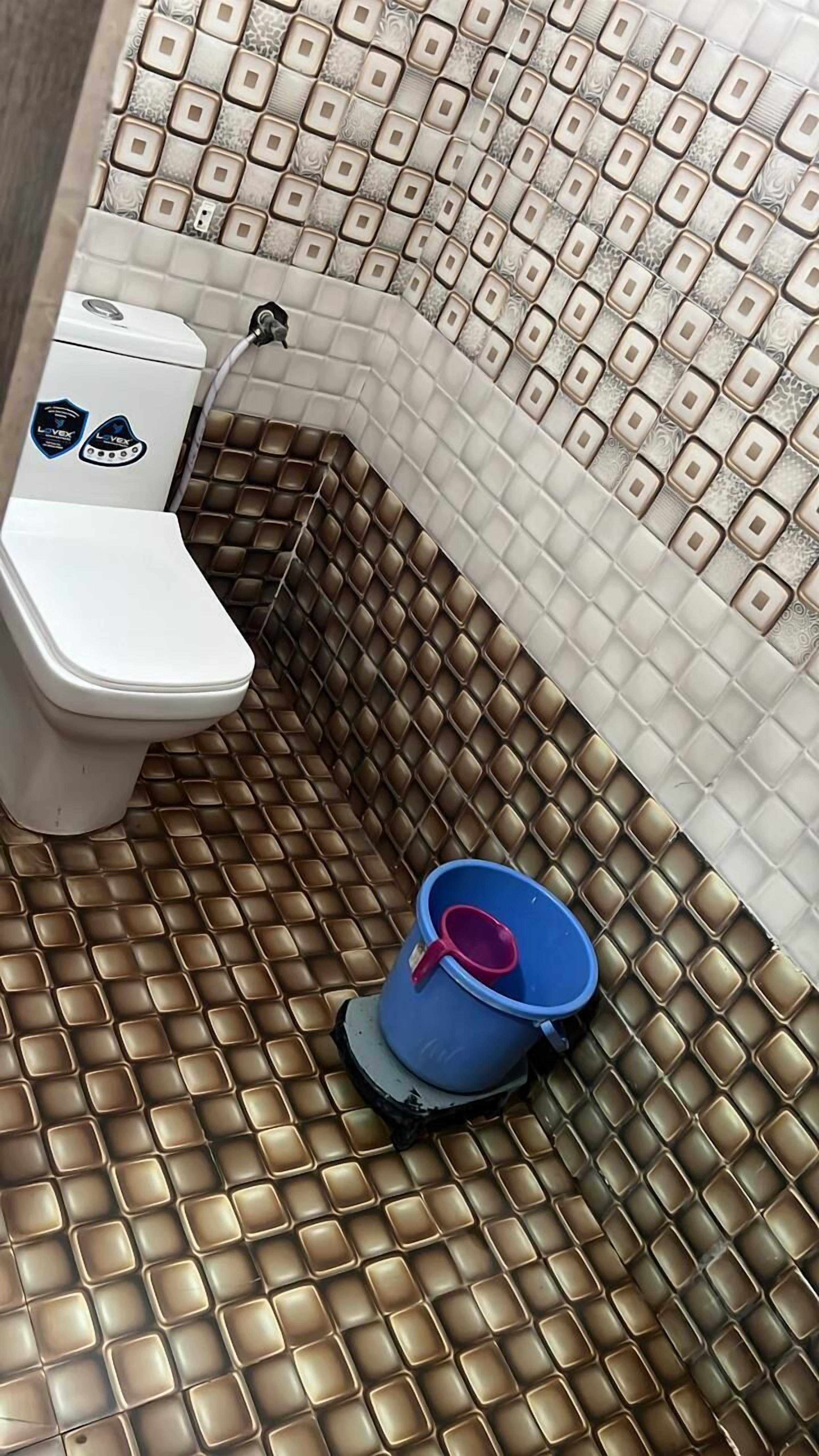 A bathroom with a toilet, a blue bucket, and a tiled floor.
