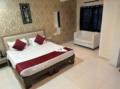 A hotel room with a large bed, a white armchair, a mirror, and a framed artwork on the wall.