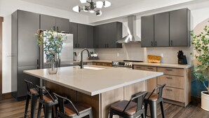 A modern kitchen with grey cabinets, a central island with bar stools, a stainless steel refrigerator, and a built-in stove. - Beautiful New Downtown Columbia Falls Condo near Glacier National Park! (Columbia Falls)