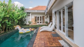 A swimming pool with a unicorn float, a wooden deck, and a house with a tiled roof and glass doors. - Villa Leya Canggu - Ella Villa Bali (Canggu)