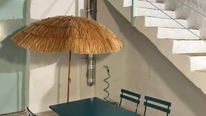 Uma área de refeições com mesa e cadeiras, um guarda-sol de palha e uma escada com degraus brancos.