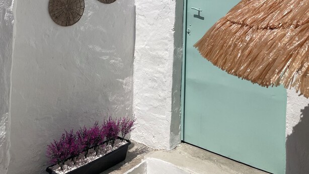 Edifício de paredes brancas com uma porta azul-turquesa, um guarda-sol de palha e um vaso com flores roxas.
