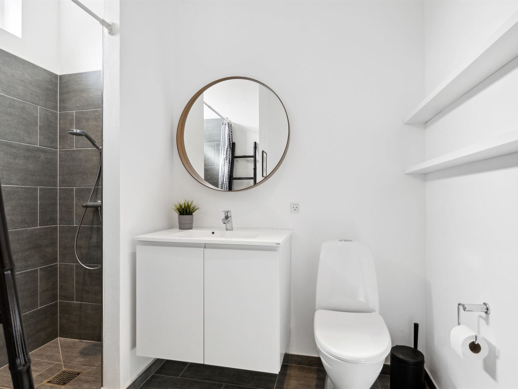 A modern bathroom with a white vanity, a toilet, a shower with dark tiles, a round mirror, and a small potted plant.
