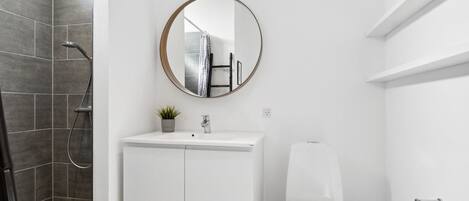 A modern bathroom with a white vanity, a toilet, a shower with dark tiles, a round mirror, and a small potted plant.