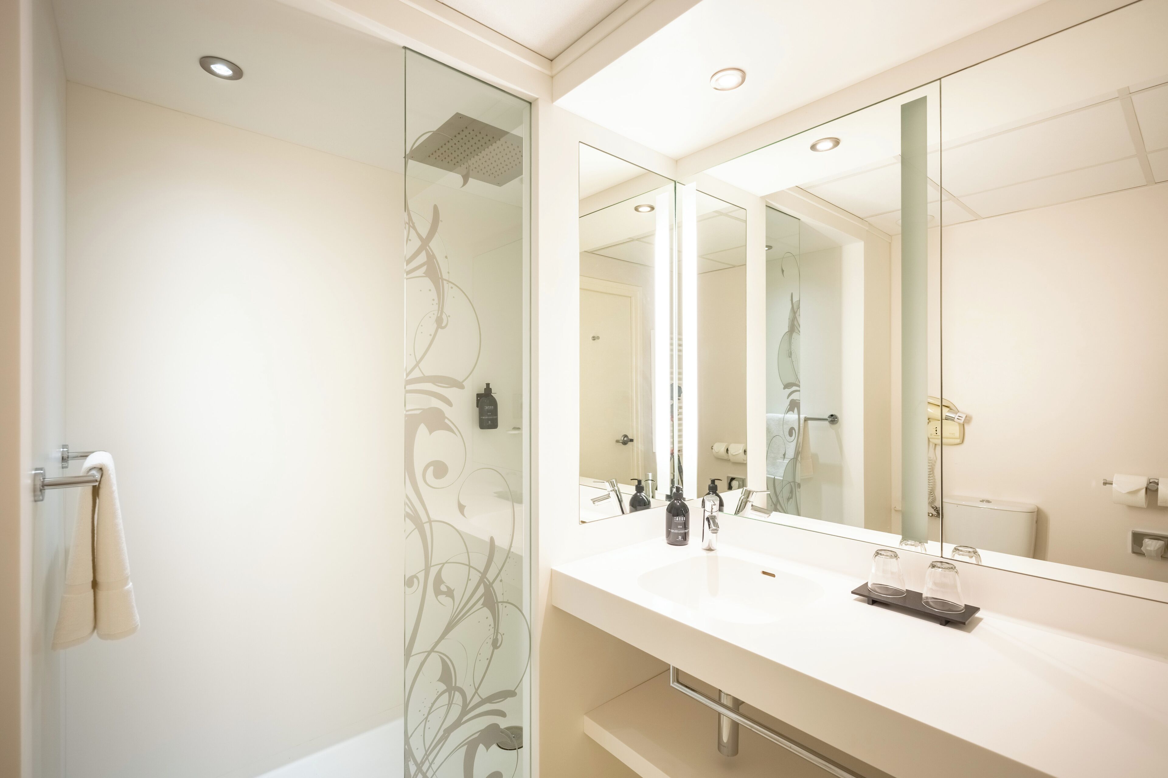 A modern bathroom with a large mirror, a glass shower enclosure, and a white sink with two glass tumblers.