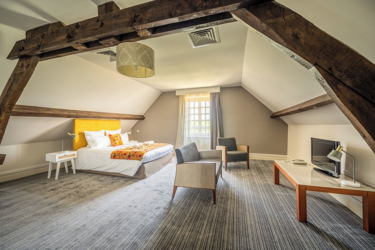 A hotel room with a loft-style ceiling, a bed with a yellow headboard, a desk with a TV, and a window with curtains.