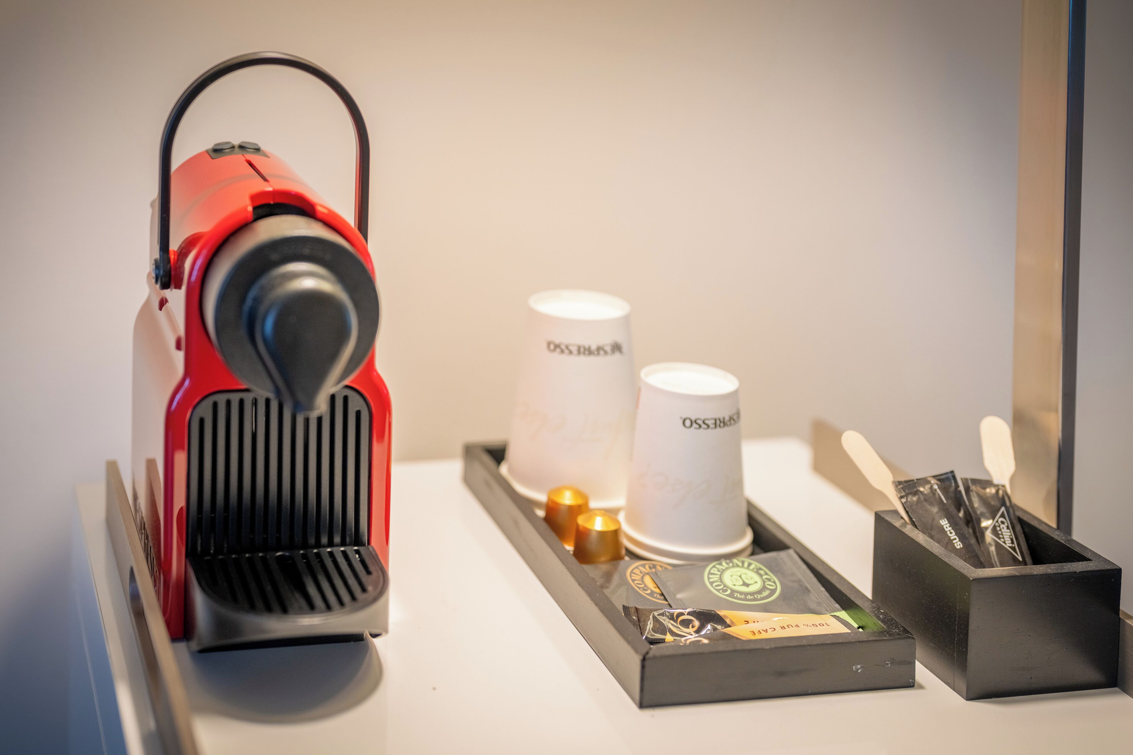 A red coffee machine on a tray with two white paper cups labeled "OSS BASSO" and a black box containing a coffee pod and utensils.