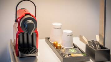 A red coffee machine on a tray with two white paper cups labeled "OSS BASSO" and a black box containing a coffee pod and utensils.