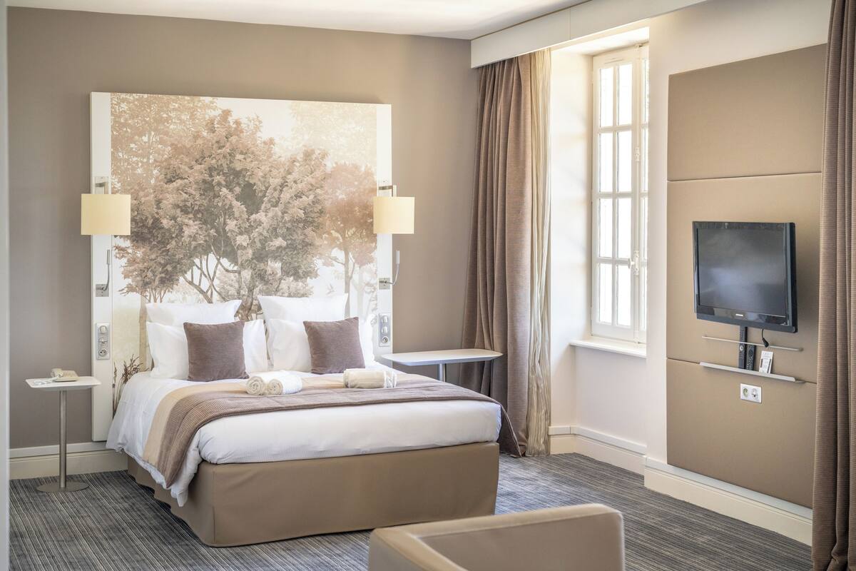 A hotel room with a large bed, a TV mounted on the wall, a small table, and a window with curtains.
