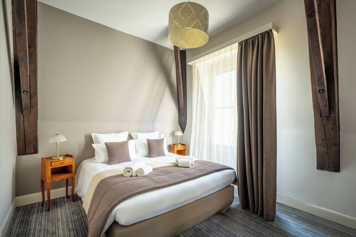 A neatly made bed with a brown headboard, a bedside table with a lamp, and a large window with curtains.