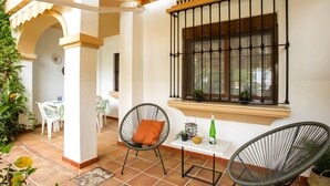 Un espace patio comprenant deux fauteuils en osier, une petite table avec une bouteille et un bol, et une fenêtre avec des barreaux en métal.