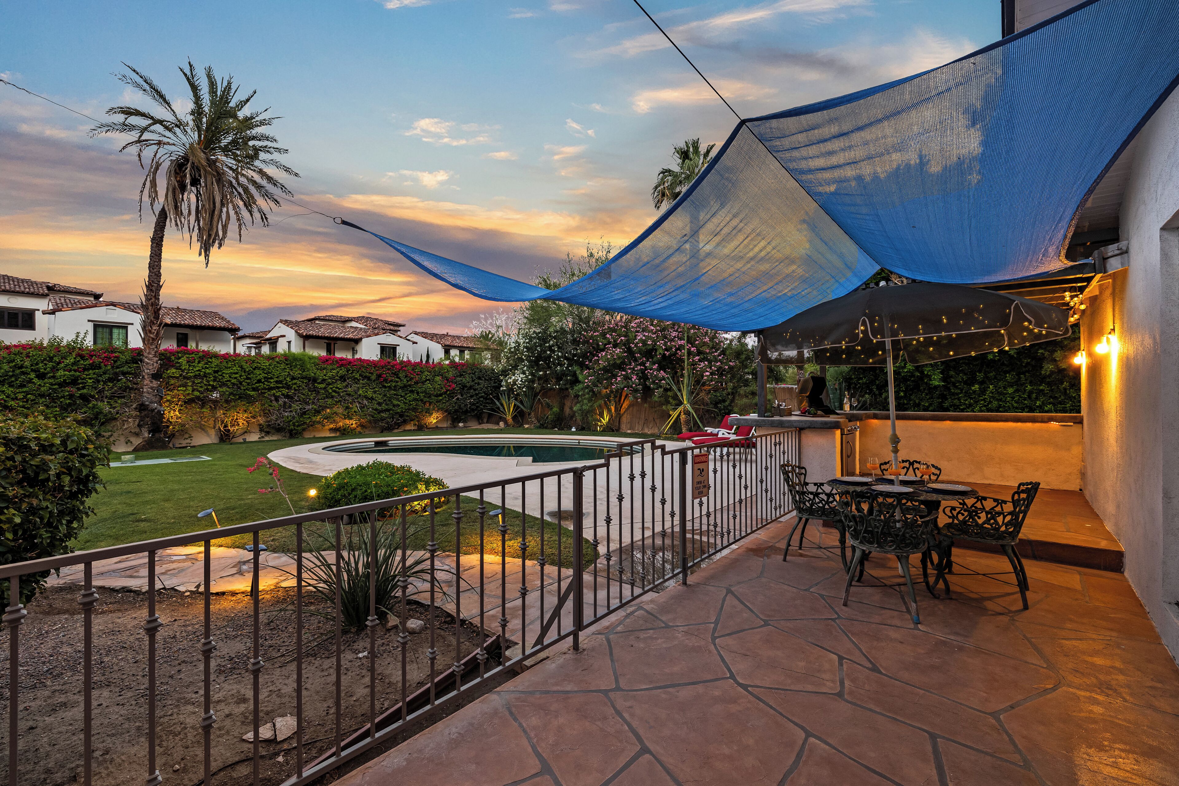 Un patio con piscina, mobiliario exterior y una vela de sombra azul.