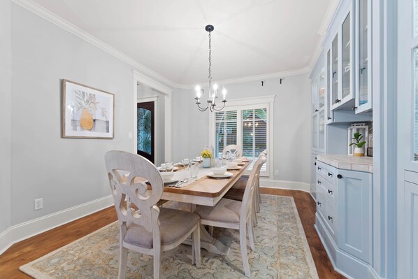 Comedor con mesa de madera dispuesta para cuatro personas, sillas blancas, una araña y armarios azul claro.